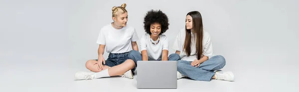 Menina americana africana alegre em t-shirt branca e jeans usando laptop perto de amigos adolescentes enquanto sentados juntos em fundo cinza, adolescentes se unindo sobre o interesse comum, banner — Fotografia de Stock