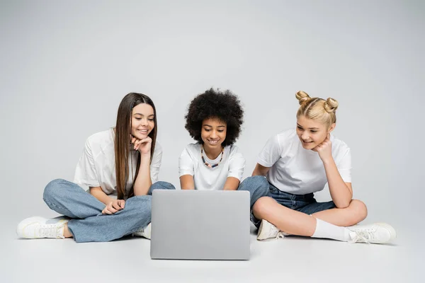 Підліток-афроамериканка використовує ноутбук біля дівчат у білих футболках і джинсах, сидячи разом на сірому фоні, підлітки зв'язуються через спільні інтереси, дружбу і спілкування. — стокове фото