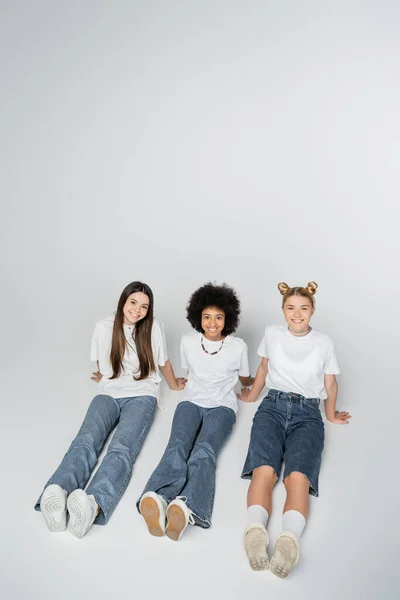 High angle view of smiling and multiethnic teenage girlfriends in white t-shirt and jeans looking at camera while sitting on grey background, adolescence models and generation z concept — Stock Photo