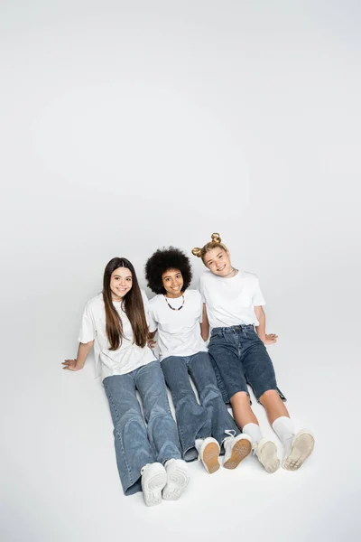 Vue en grand angle d'amis adolescents positifs et multiethniques en t-shirts et jeans blancs regardant la caméra et assis ensemble sur fond gris, les modèles d'adolescence et le concept de génération z — Photo de stock