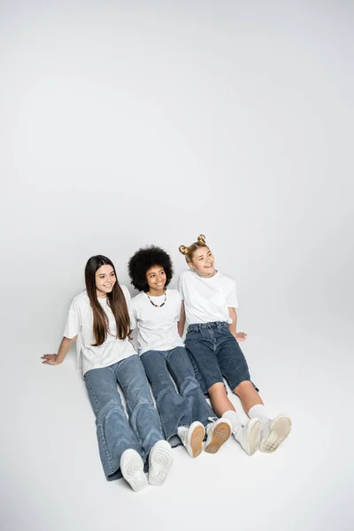 Blickwinkel auf positive interrassische Teenager-Freundinnen in weißen T-Shirts und blauen Jeans, die wegschauen, während sie auf grauem Hintergrund sitzen, Adoleszenz-Models und Generation-Z-Konzept — Stockfoto