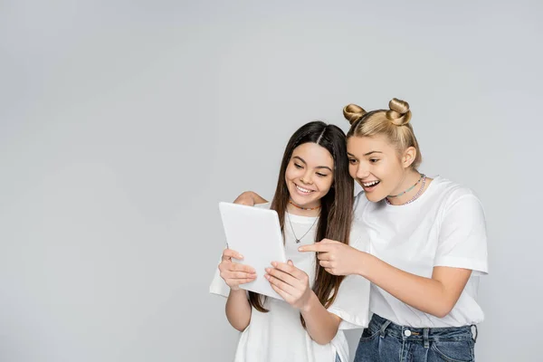 Smiling blonde teen girl in jeans and white t-shirt pointing with finger at digital tablet and hugging brunette friend isolated on grey, teenagers bonding over common interest, Touch Screen — Stock Photo