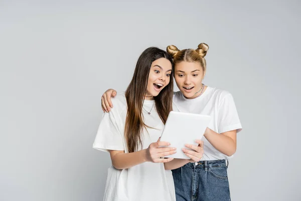 Copines adolescentes excitées en t-shirts blancs utilisant la tablette numérique ensemble tout en étant isolé sur gris, les adolescents collant sur l'intérêt commun, l'amitié et la compagnie — Photo de stock
