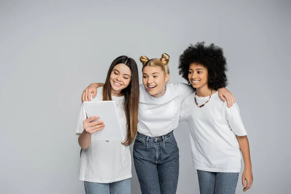 Adolescent blond positif en t-shirt blanc et en jeans embrassant des copines multiethniques et utilisant une tablette numérique tout en étant isolé sur du gris, les adolescents se lient d'intérêt commun — Photo de stock