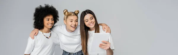 Smiling interracial teenage girlfriends in white t-shirts using digital tablet together while hugging and standing isolated on grey, teenagers bonding over common interest, banner — Stock Photo
