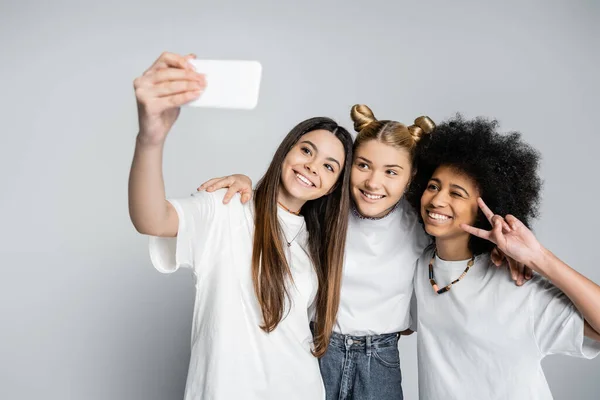 Усміхнені дівчата-підлітки в білих футболках обіймаються і жестикулюють, беручи селфі на смартфон на сірому фоні, підлітки зв'язуються над спільними інтересами, дружбою і товариством — стокове фото