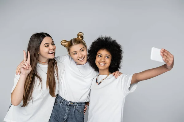 Усмішка підліткової афроамериканської дівчини в білій футболці вживає сельфі на смартфоні з подругами, які обіймають один одного ізольовано на сірому, підлітки зв'язуються через спільні інтереси, дружбу. — стокове фото