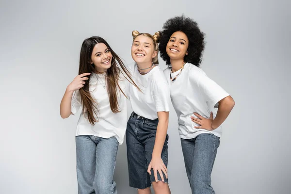 Petites amies adolescentes élégantes et joyeuses en jeans et t-shirts blancs regardant la caméra tout en posant ensemble sur fond gris, modèles d'adolescence et concept de génération z — Photo de stock