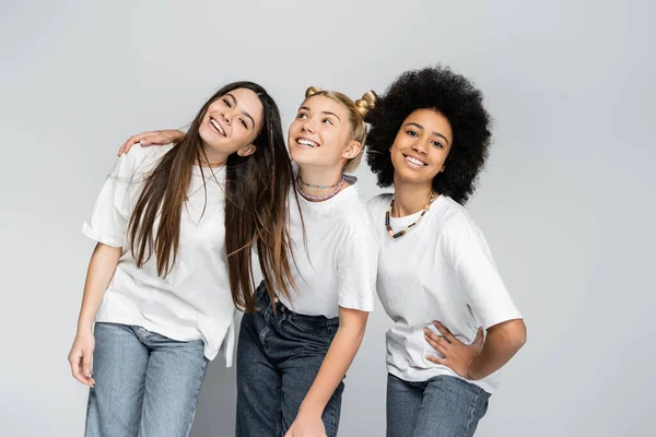 Novias adolescentes multiétnicas en jeans y camisetas blancas casuales posando mientras se abrazan y se paran juntas aisladas en modelos grises, adolescentes y concepto de generación z, amistad - foto de stock