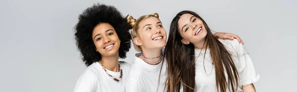 Positive and multiethnic teenage girlfriends in casual white t-shirts hugging and looking at camera while standing isolated on grey, adolescence models and generation z concept, banner — Stock Photo