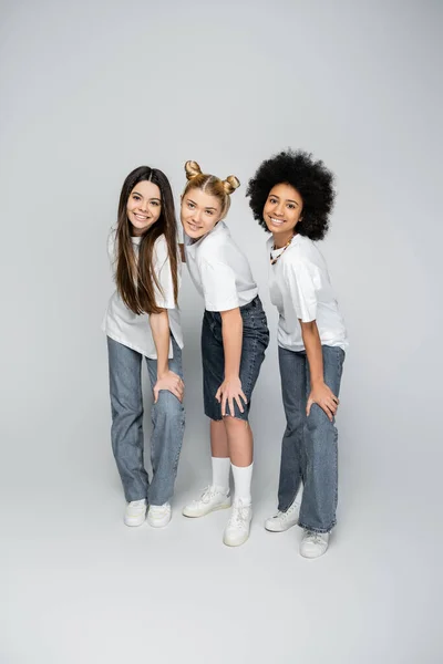 Volle Länge stilvolle und jugendlich multiethnische Freundinnen in lässigen weißen T-Shirts, Jeans und Turnschuhen, die gemeinsam posieren, während sie auf grauem Hintergrund stehen, Adoleszenz-Models und Generation-Z-Konzept — Stockfoto