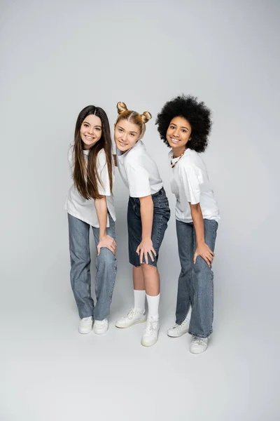 Longitud completa de novias multiétnicas elegantes y adolescentes en camisetas blancas casuales, jeans y zapatillas de deporte posando juntas mientras están de pie sobre un fondo gris, modelos de adolescencia y concepto de generación z - foto de stock