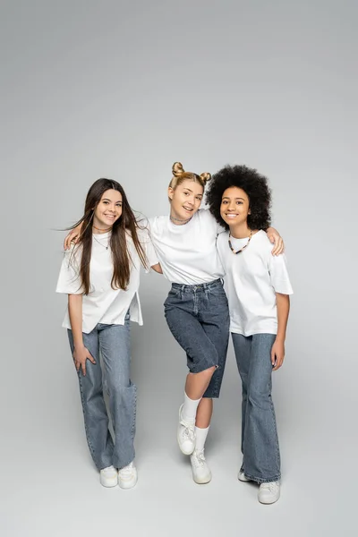 Full length of joyful blonde teen girl looking at camera while hugging multiethnic friends in white t-shirts and jeans and standing on grey background, adolescence models and generation z concept — Stock Photo