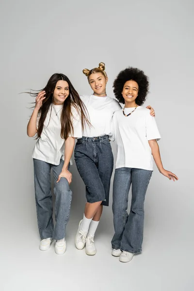 Повна довжина стильної дівчини-підлітка обіймає багатоетнічних і усміхнених подруг у білих футболках і джинсах, стоячи на сірому фоні, підліткових моделях і концепції покоління z — стокове фото