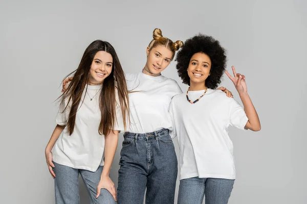 Модна блондинка дівчина-підліток в джинсах і білій футболці обіймає багатоетнічних подруг і позує разом ізольовано на сірих, підліткових моделях і концепції покоління z, дружби і компаньйона — стокове фото