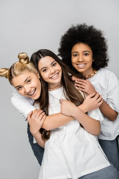 Portrait of cheerful and teen multiethnic girlfriends in jeans and white t-shirts hugging brunette friend while posing isolated on grey, adolescence models and generation z concept — Stock Photo
