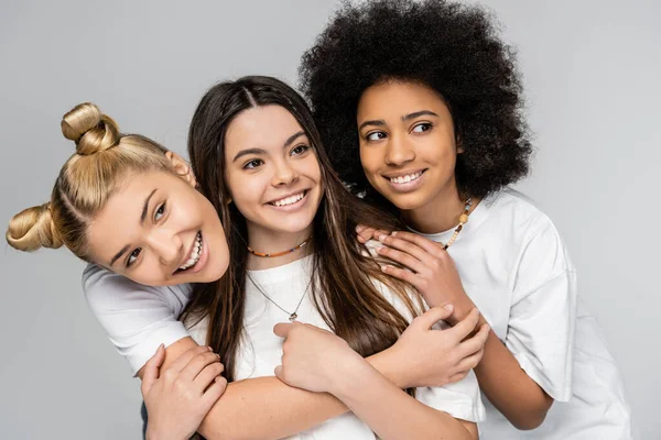 Porträt fröhlicher und multiethnischer Teenager in weißen T-Shirts, die sich umarmen und wegschauen, während sie isoliert auf grauen, pubertären Mädchen und dem Konzept der Generation Z, Freundschaft und Kameradschaft posieren. — Stockfoto
