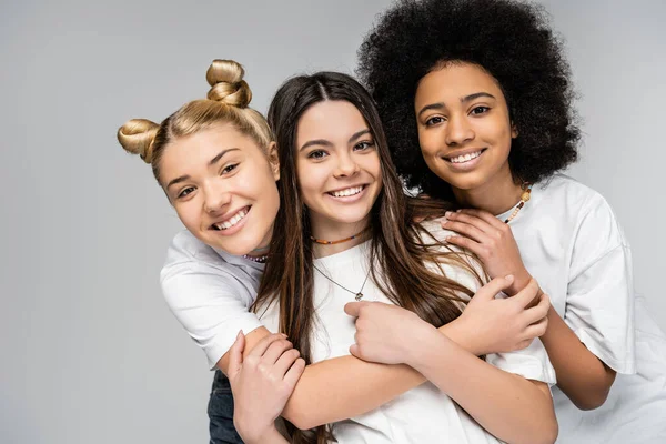Portrait of cheerful and multiethnic teenage girls in white t-shirts hugging each other and looking at camera while standing isolated on grey, teenage friends having fun together — Stock Photo