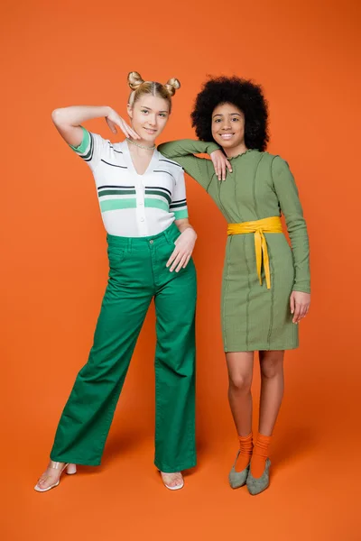 Full length of stylish and multiethnic teen girlfriends in trendy outfits posing and smiling at camera while standing together on orange background, trendy generation z concept — Stock Photo