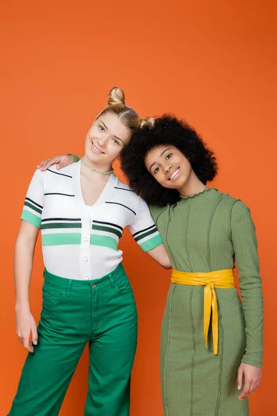 Smiling and trendy blonde teenage girl in trendy outfit hugging african american girlfriend and looking at camera while standing isolated on orange, trendy generation z concept — Stock Photo