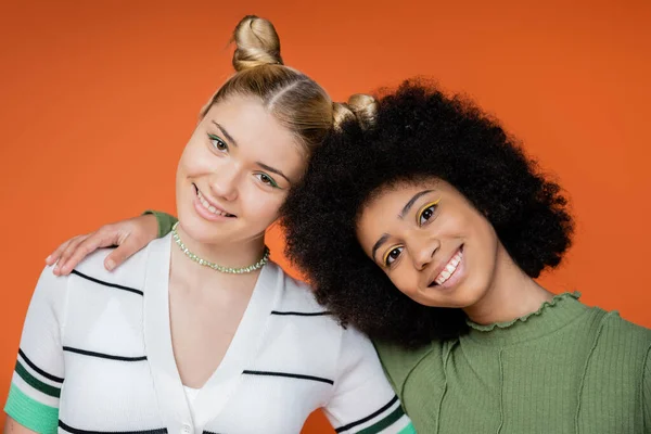 Porträt eines stilvollen und positiven afrikanisch-amerikanischen Mädchens, das in die Kamera blickt, während es seine blonde Teenie-Freundin umarmt und isoliert auf einem orangefarbenen, trendigen Konzept der Generation Z steht — Stockfoto