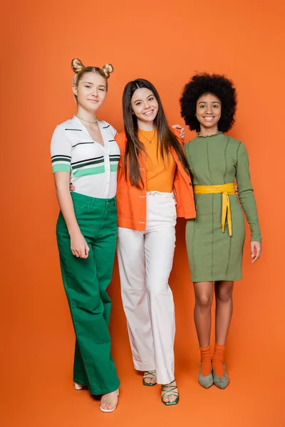 Полная длина стильных и многонациональных девочек-подростков в модных нарядах, обнимающихся, позируя и глядя на камеру вместе в оранжевом фоне, подростковой моды, подростков моделей — стоковое фото