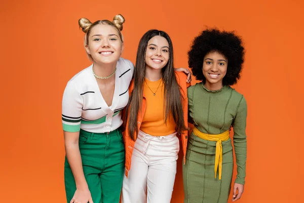 Positive und multiethnische Teenager-Freundinnen in trendigen Outfits, die sich umarmen und vor orangefarbenem Hintergrund in die Kamera lächeln, trendiges Generation-Z-Konzept, Freundschaft und Kameradschaft — Stockfoto