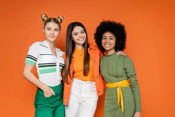 Smiling brunette teen girl in trendy outfit hugging stylish multiethnic girlfriends while posing together and standing on orange background, trendy generation z concept, friendship and companionship — Stock Photo