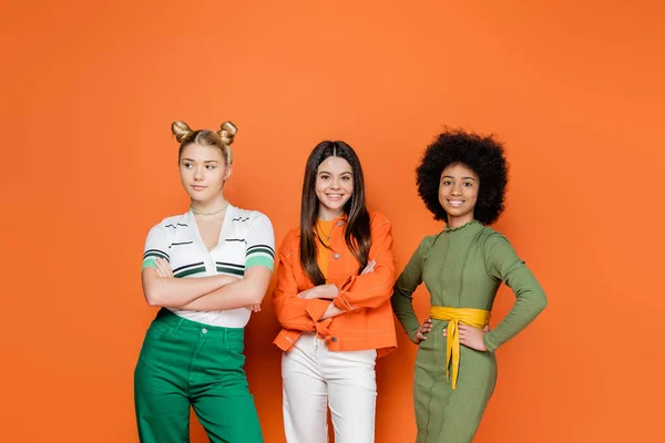 Positive and interracial teenage friends in trendy outfits crossing arms while standing and posing together on orange background, trendy generation z concept, friendship and companionship — Stock Photo