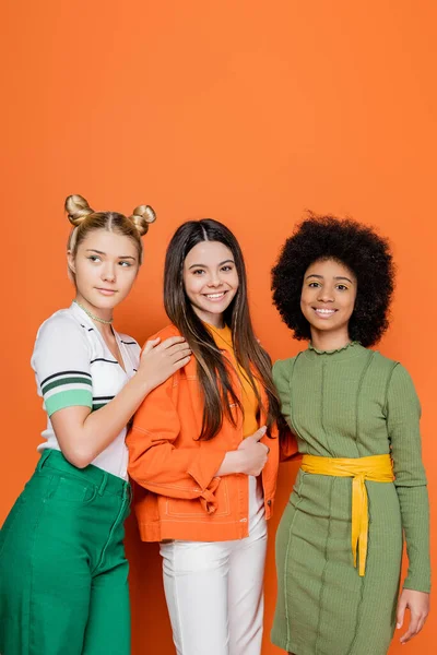 Smiling brunette teen girl in stylish outfit looking at camera near multiethnic girlfriends while posing together on orange background, trendy generation z concept, friendship and companionship — Stock Photo