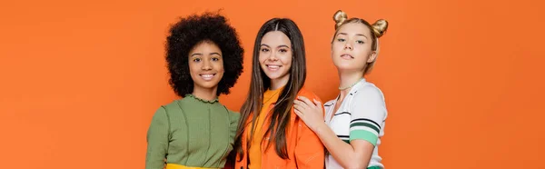 Trendy blonde teenage girl hugging multiethnic friends in stylish outfits while standing and posing together isolated on orange, trendy generation z concept, banner, friendship and companionship — Stock Photo