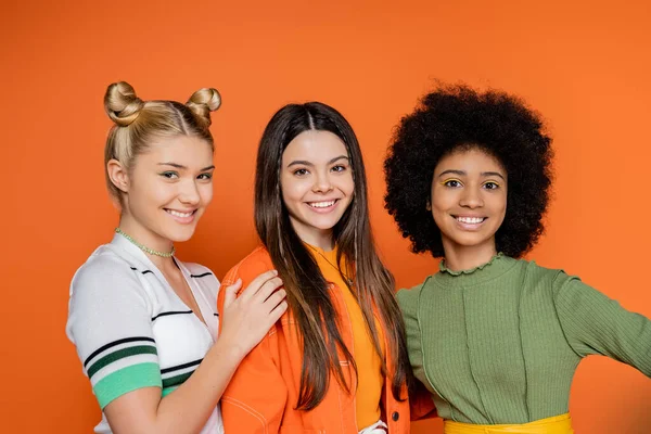 Portrait of positive and multiethnic teenage girlfriends in stylish outfits with makeup looking at camera while standing isolated on orange, trendy generation z concept, friendship and companionship — Stock Photo