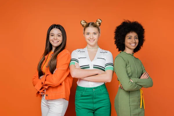 Adolescentes multiétnicos confiados y de moda cruzando brazos mientras posan juntos sobre fondo naranja, concepto de generación z de moda, amistad y compañía - foto de stock