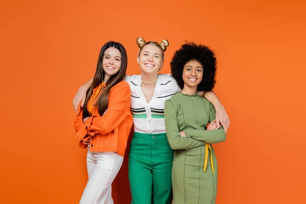 Cheerful blonde teenage girl hugging fashionable multiethnic girlfriends in trendy outfits crossing arms and looking at camera on orange background, trendy generation z concept — Stock Photo