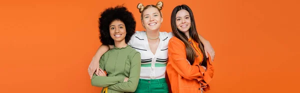 Confident and smiling blonde teenager hugging stylish and multiethnic girlfriends crossing arms and looking at camera isolated on orange, trendy generation z concept, banner, friendship — Stock Photo