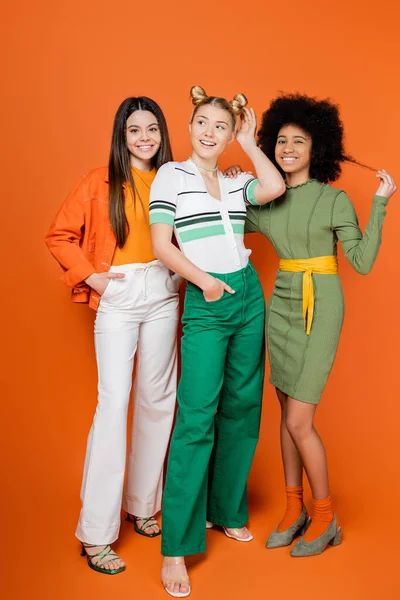 Full length of cheerful and confident multiethnic teen girlfriends in trendy outfits posing while standing together on orange background, cultural diversity and generation z fashion concept — Stock Photo