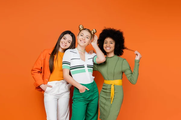Multiethnic and teenage girlfriends in trendy outfits with makeup posing together and looking at camera on orange background, cultural diversity and generation z fashion concept, friendship — Stock Photo