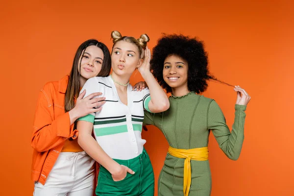 Trendy and multiethnic girlfriends hugging stylish blonde friend pouting lips while posing together on orange background, cultural diversity and generation z fashion concept, friendship — Stock Photo
