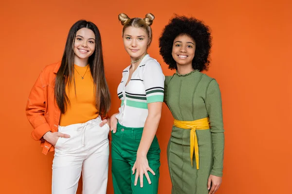 Stylish multiethnic teenage girlfriends in trendy outfits posing together and looking at camera while standing on orange background, cultural diversity and generation z fashion concept — Stock Photo