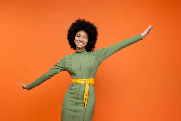 Cheerful and teen african american girl in stylish green dress and bold makeup winking and looking at camera while standing isolated on orange, teenage fashion and generation z concept — Stock Photo