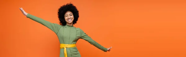 Smiling african american teen girl with bold makeup wearing green dress and winking at camera isolated on orange, teenage fashion and generation z concept, banner with copy space — Stock Photo
