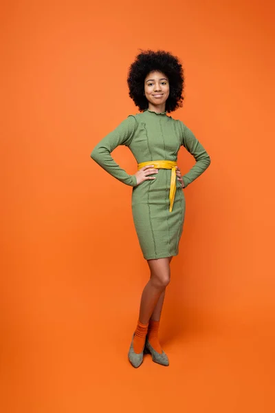 Full length of smiling and fashionable african american teen girl with bold makeup posing in green dress while standing on orange background, teenage fashion and generation z concept — Stock Photo