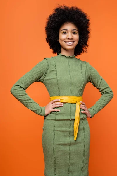 Stylish and teen african american girl with bold makeup wearing green dress and holding hands on hips while standing isolated on orange, teenage fashion and generation z concept — Stock Photo