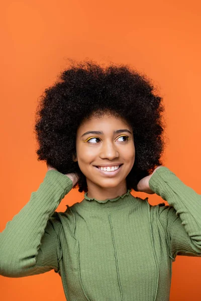 Porträt eines fröhlichen afrikanisch-amerikanischen Mädchens im Teenageralter mit kühnem Make-up und grünem Kleid, das die Haare berührt und isoliert auf orangefarbene Teenagermode und das Konzept der Generation Z blickt — Stockfoto
