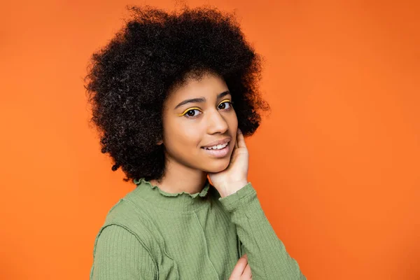 Positive and stylish african american teen girl with bright makeup and green dress touching cheek and looking at camera isolated on orange, teenage fashion and generation z concept — Stock Photo