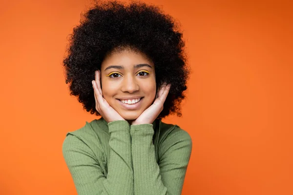 Portrait of happy teen african amerian girl with bold makeup wearing green dress and touching cheeks while standing isolated on orange, youth culture and generation z concept — Stock Photo