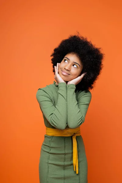 Dreamy and stylish teen african american girl with bold makeup wearing green dress and touching cheeks while standing isolated on orange, youth culture and generation z concept — Stock Photo