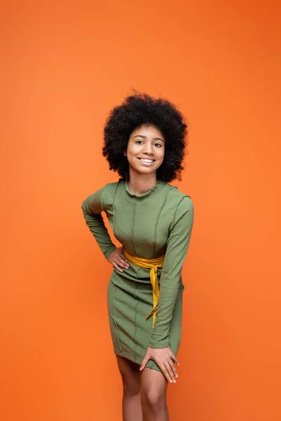 Трендовая и позитивная африканская девочка-подросток с ярким макияжем в зеленом платье и держащаяся за бедро, изолированная от оранжевого, молодежная культура и концепция поколения Z — стоковое фото