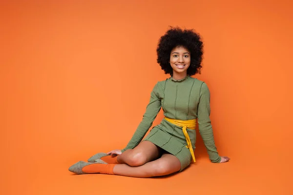 Joyeux et adolescent afro-américaine en robe verte à la mode souriant à la caméra tout en étant assis et posant sur fond orange, la culture de la jeunesse et la génération z concept — Photo de stock