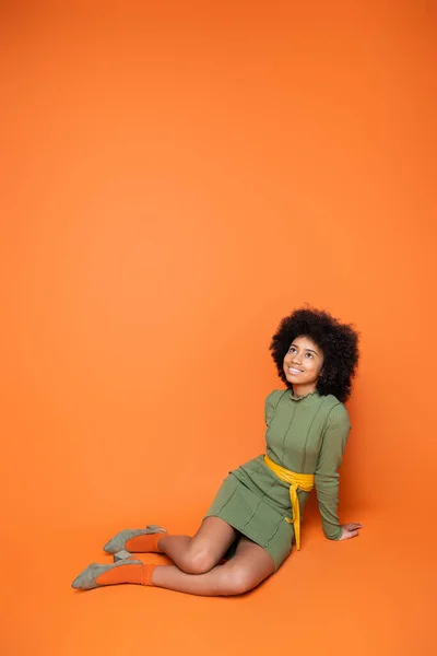 Volle Länge des fröhlichen und trendigen afrikanisch-amerikanischen Teenagers in grünem Kleid, der aufschaut, während er auf orangefarbenem Hintergrund sitzt und posiert, Jugendkultur und Generation-Z-Konzept — Stockfoto